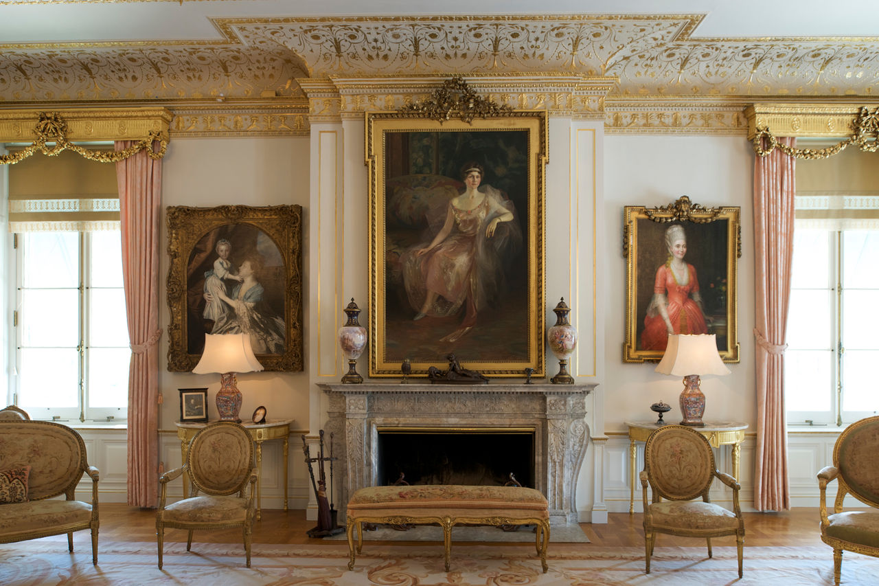A large living room with a stone fireplace and three women's painted portraits surrounded by elegant pink decor and furniture for sitting. 